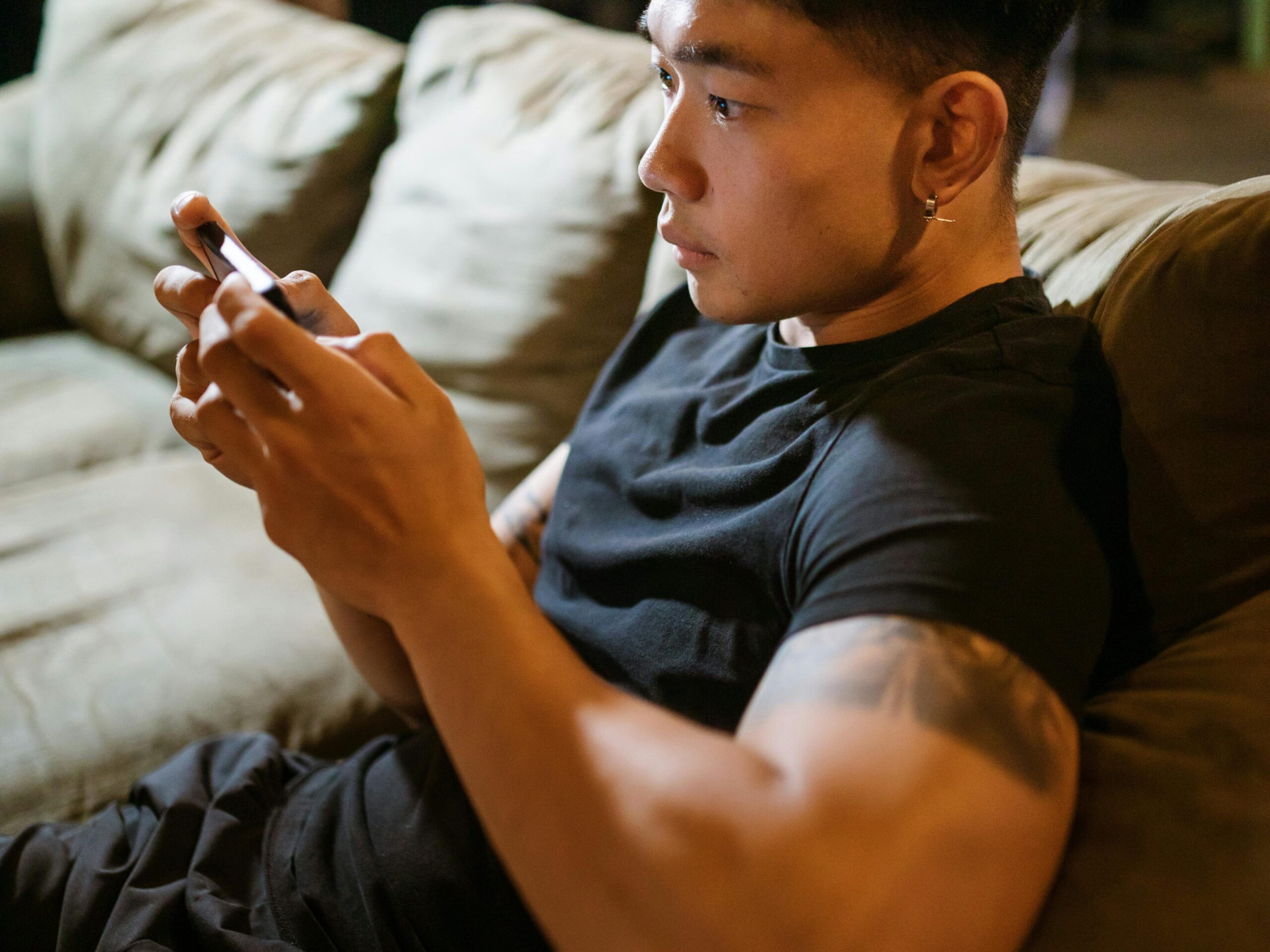 A young man intensely focused on playing a game on his smartphone while lounging on a couch indoors.